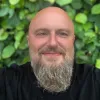 Smiling bald man with a full beard wearing a black shirt standing in front of green leafy background.